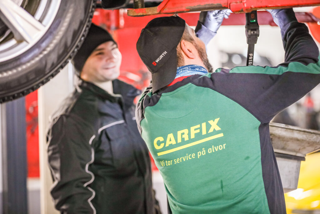 Bilde av Carfix-mekaniker som står under en bil som står på løftebukk. Han holder på å skru noe fast under bilen. En kollega står smilende og ser på.