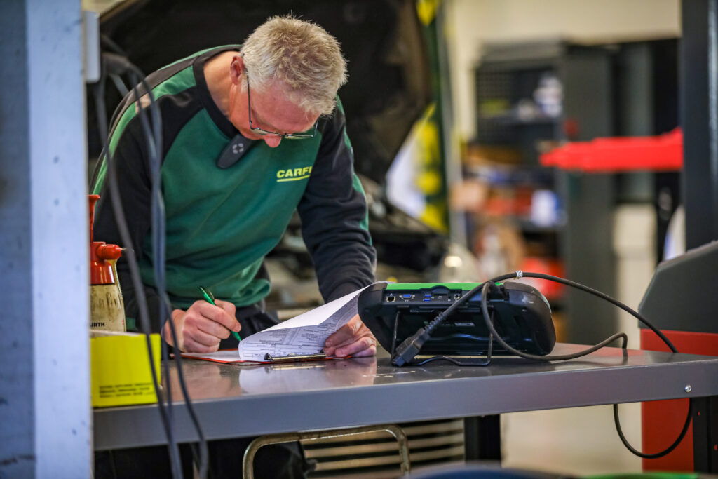Bilde av mekaniker som står i verkstedet, bøyd over et kontrollskjema som han noterer på. Han har utført EU-kontroll på dagen på bilen i bakgrunnen. Carfix utfører også etterkontroll.