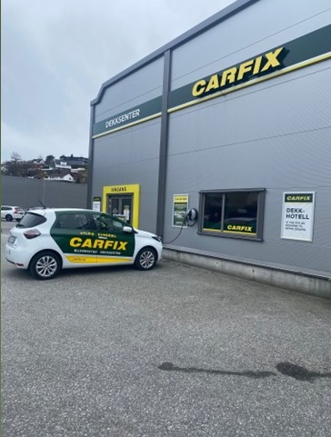 Bilde av utsiden av Carfix Loddefjord med Carfix-profilering. Inngangen til kundemottaket er helt til venstre på bygget, og utenfor står lånebilen i Carfix-profilering, tilkoblet ladeboks.