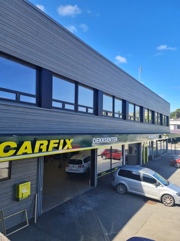 Bilde av utsiden av Carfix Gann, med Carfix-profilering. Bildet viser 2 åpne verkstedsporter og nøkkelautomatboks hengende på veggen til venstre. Plassen utenfor er asfaltert.