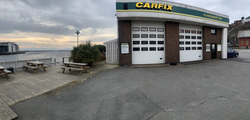 Bilde av utsiden av Carfix Øyro med Carfix-profilering. Det er to verkstedsporter og inngang til kundemottaket til høyre. På venstre siden ser man utløpet av Oselven, og bryggen der det står noen benker.