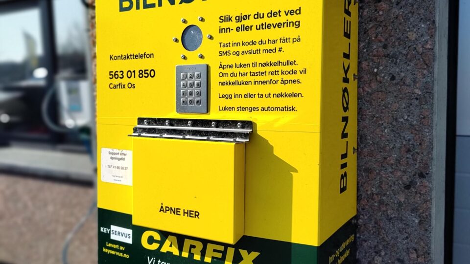 Bilde av automatisk nøkkelautomat som henger på murvegg utenfor et Carfix-verksted. Boksen er gul, og det står "bilnøkler" med stor skrift øverst på boksen. Vi har nøkkeldropp!